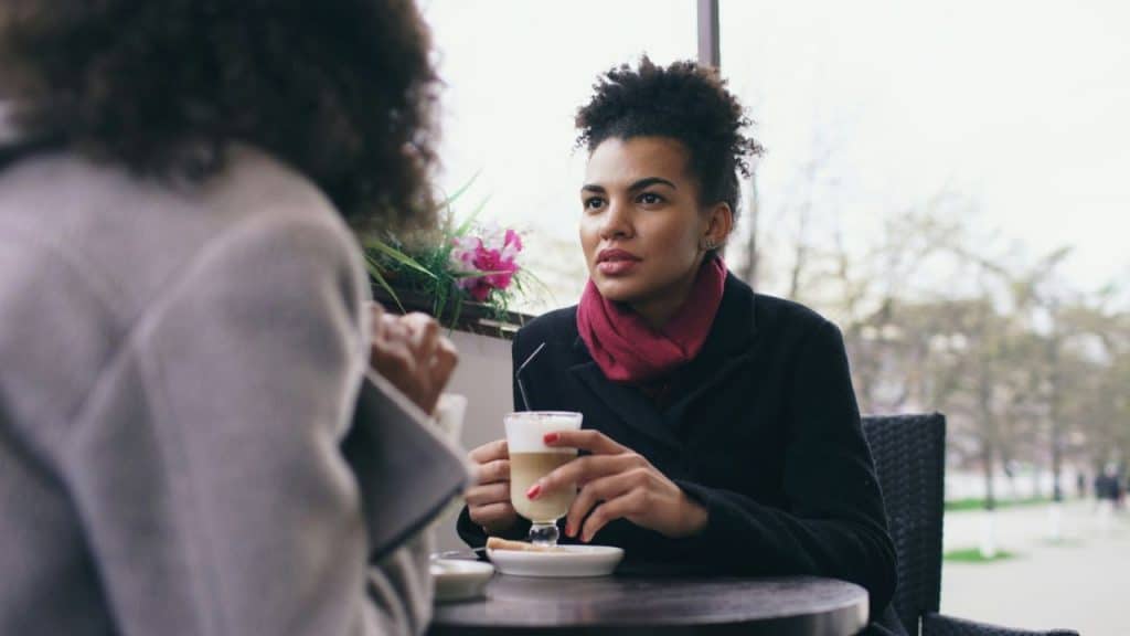 A woman with curly hair wearing a red scarf sits at a cafe holding a latte.