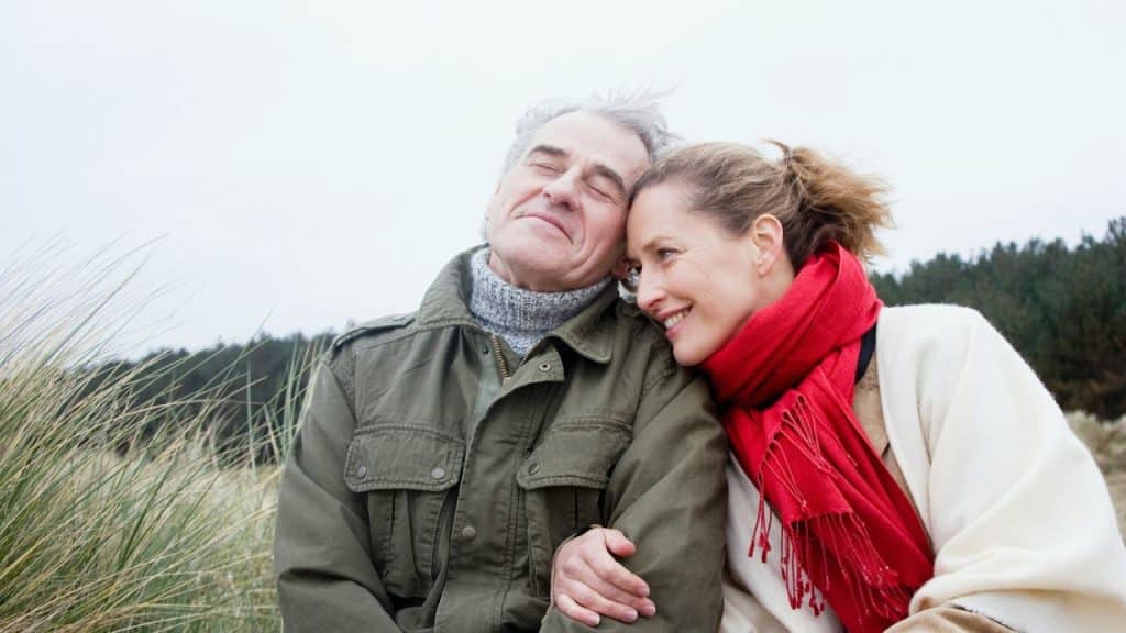 An older man and woman sit together outdoors, leaning their heads against each other.