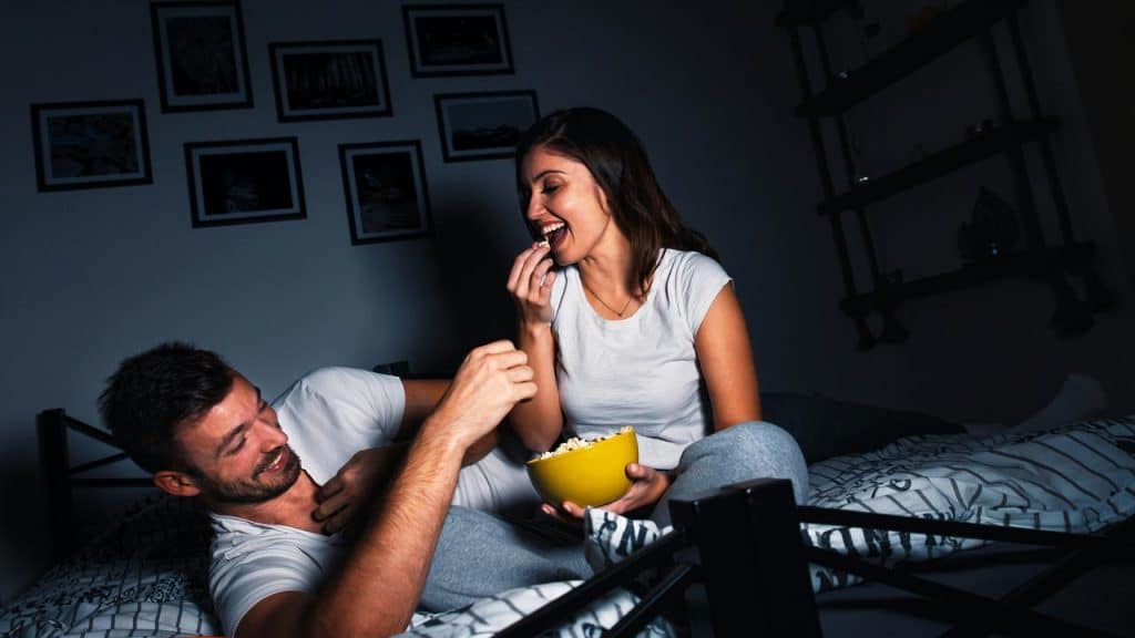 A couple relaxing on a bed while sharing popcorn and laughing together.
