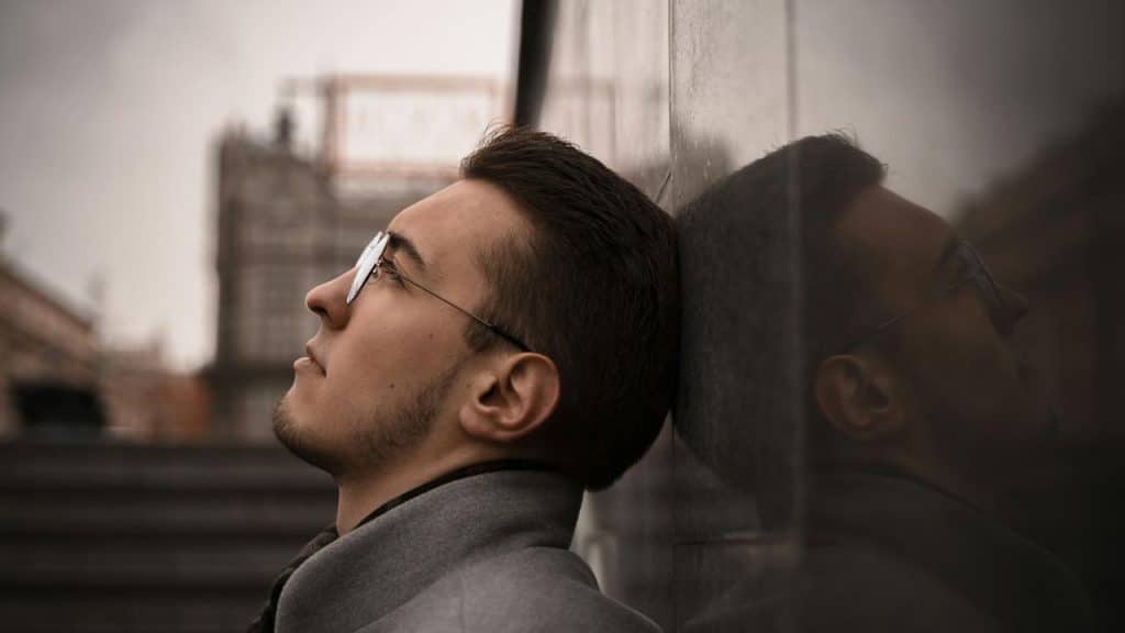 A man in glasses leaning his head back against a reflective wall outdoors.
