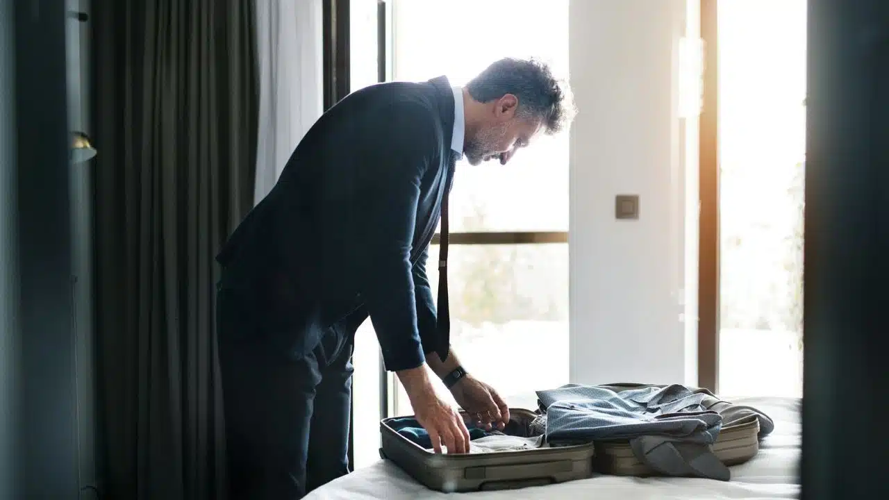 A man in a suit leans over a bed to pack items into a suitcase.