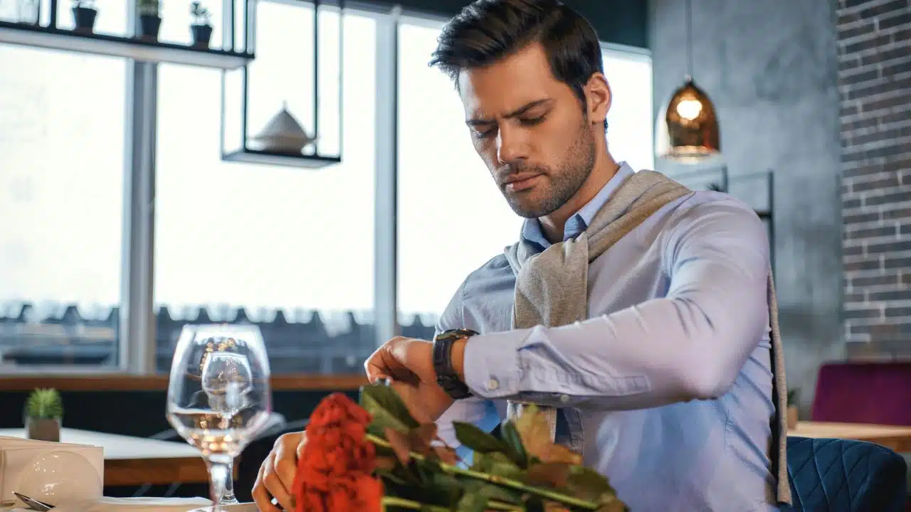 A man in a light blue shirt checks his watch while sitting near red roses.
