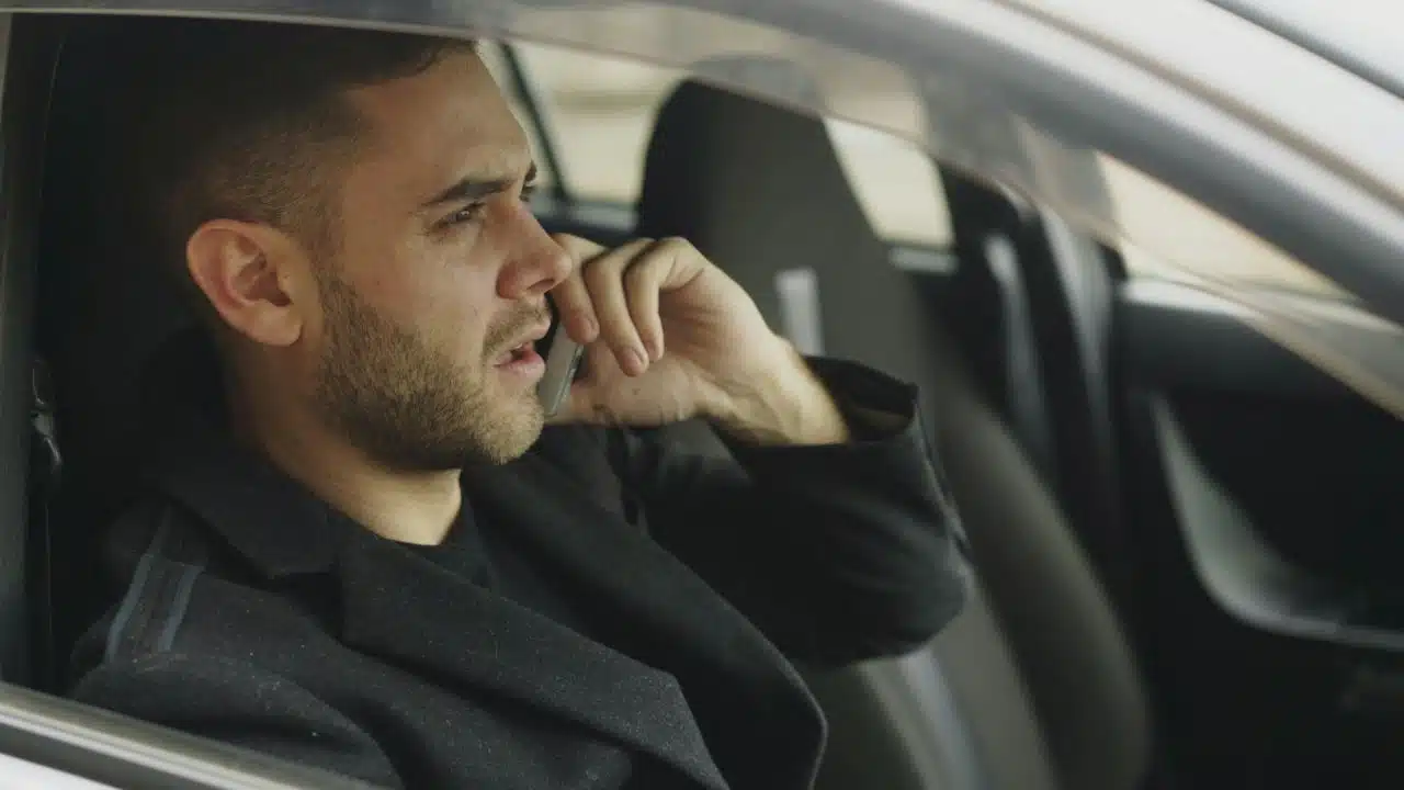 A bearded man wearing a dark coat sits in a car while talking on a cellphone.