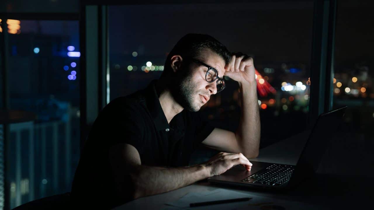 A man wearing glasses works on a laptop in a dark room at night.