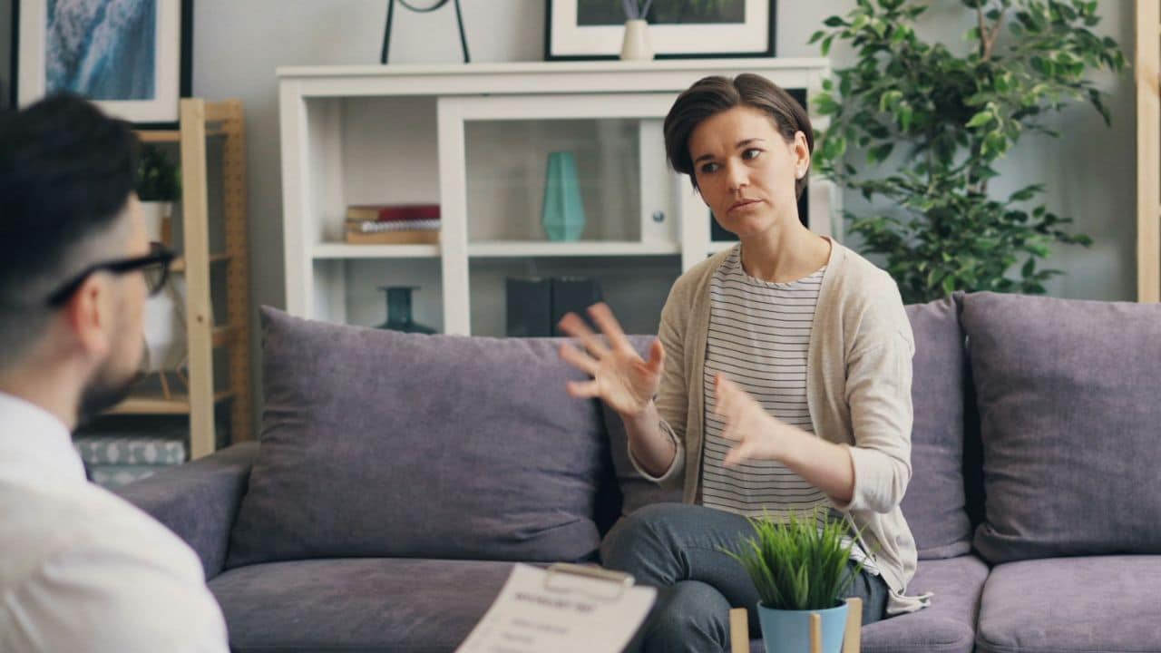 A woman sits on a purple couch and gestures while talking to a man.