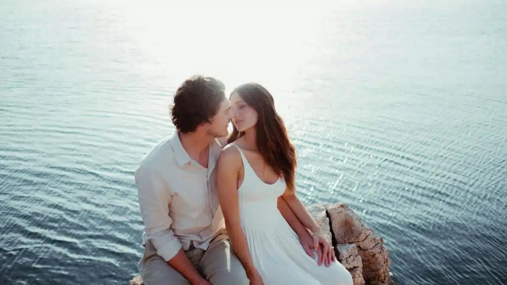 A man and a woman sitting on a rock by the water.