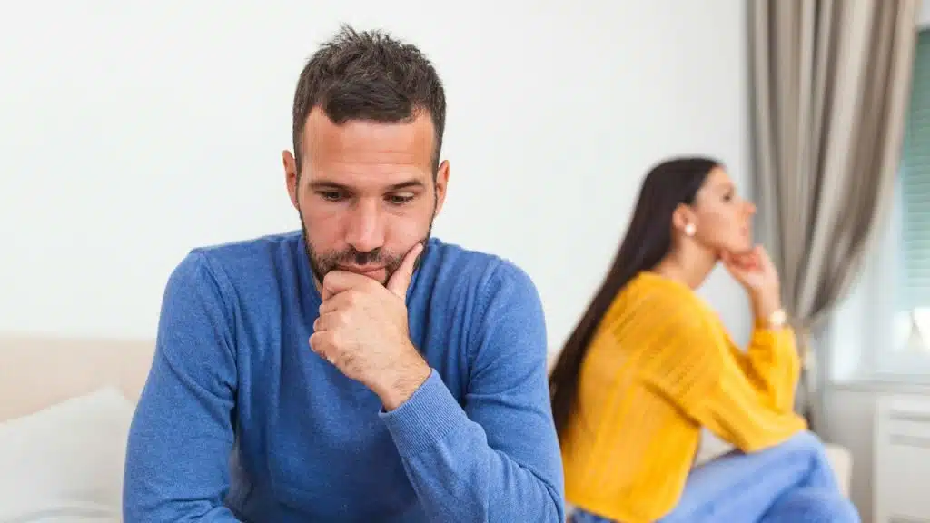 A man looking forlorn as a woman sits with an inscrutable expression in the background.