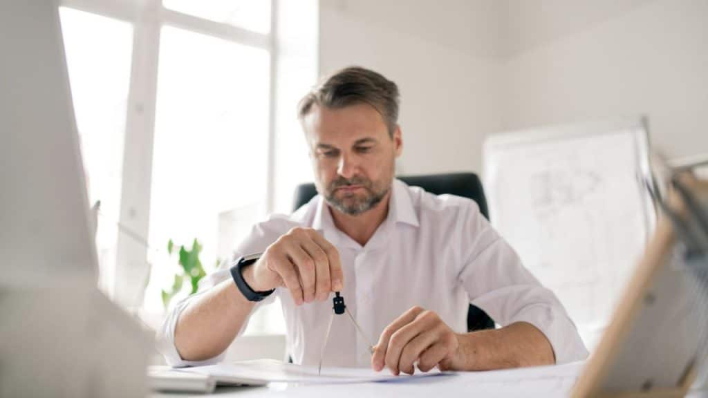 A man in a white shirt sits at a desk using a drawing compass on paper.