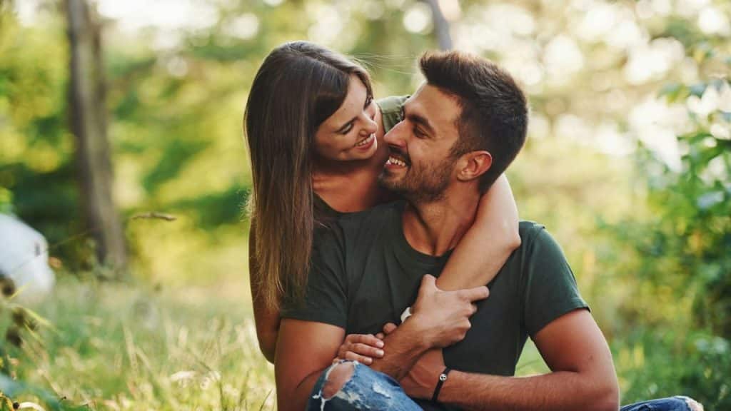 A woman with long brown hair hugs a man from behind while they sit outdoors.
