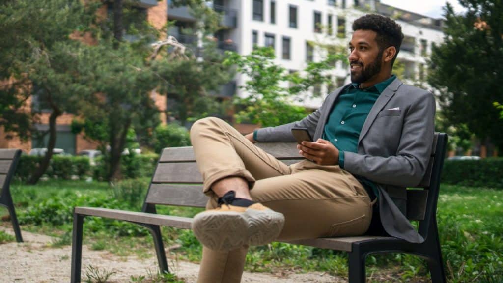A smiling man in a grey blazer sits on a park bench holding a smartphone.