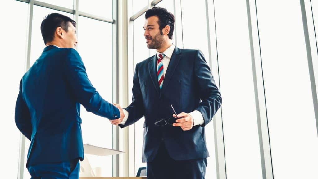 Two men in business suits smile while shaking hands in a bright, modern office building.