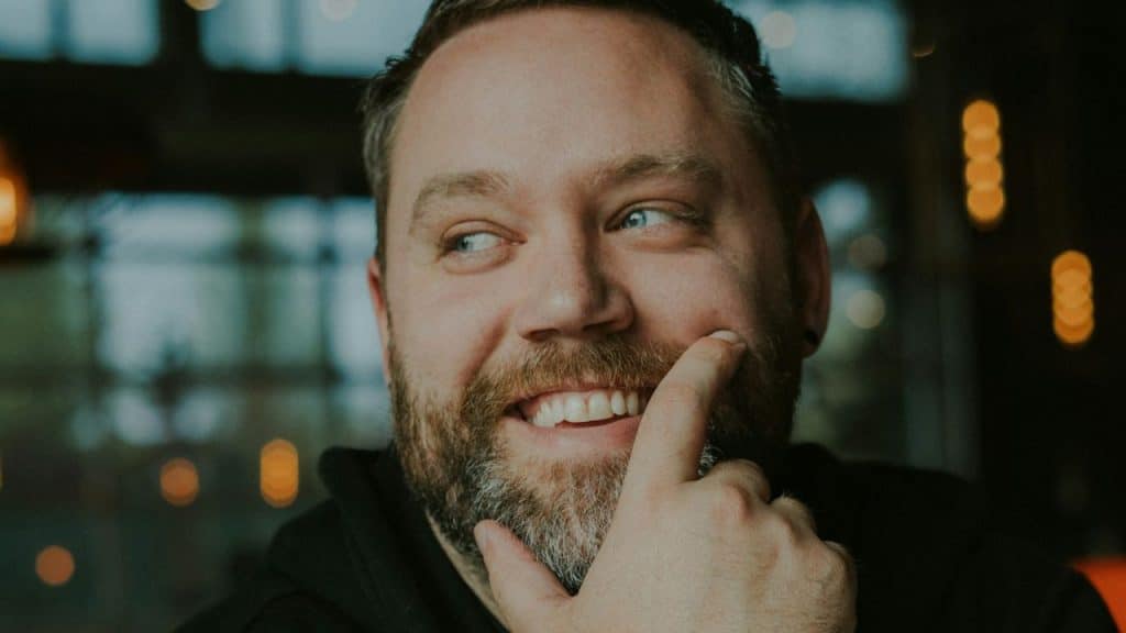 A man with a beard smiles and looks to the side with a finger on his cheek.