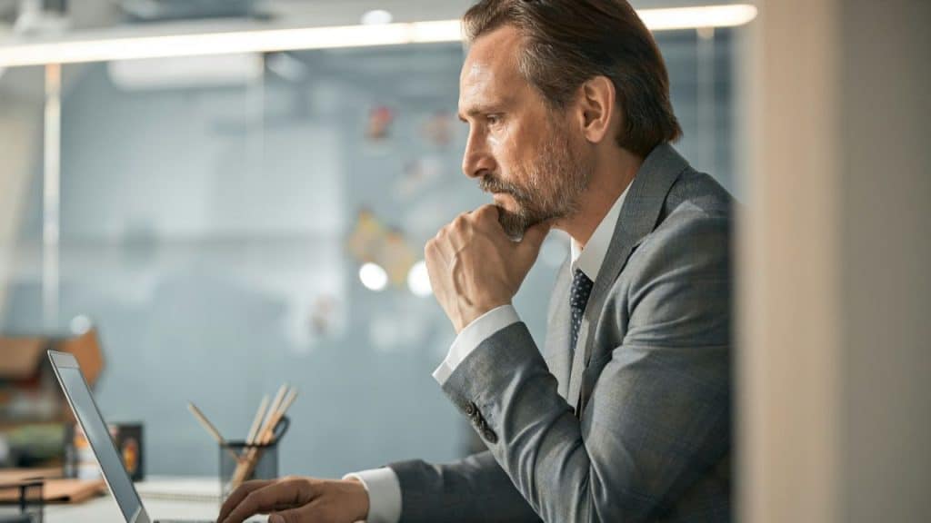 A man in a gray suit rests his chin on his hand while looking at a laptop.