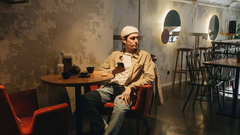 A man in a beanie sits at a cafe table holding a phone and looking away.