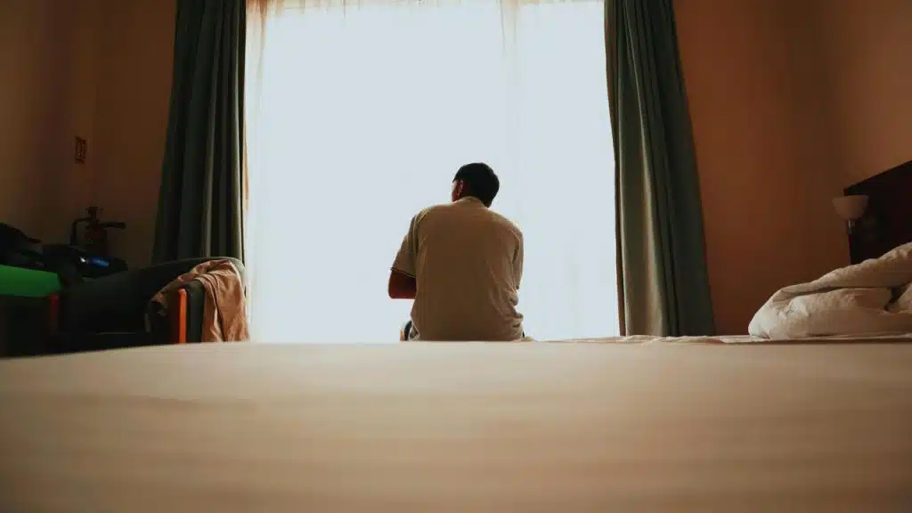 A person sitting on a bed, facing a bright window in a softly lit room.