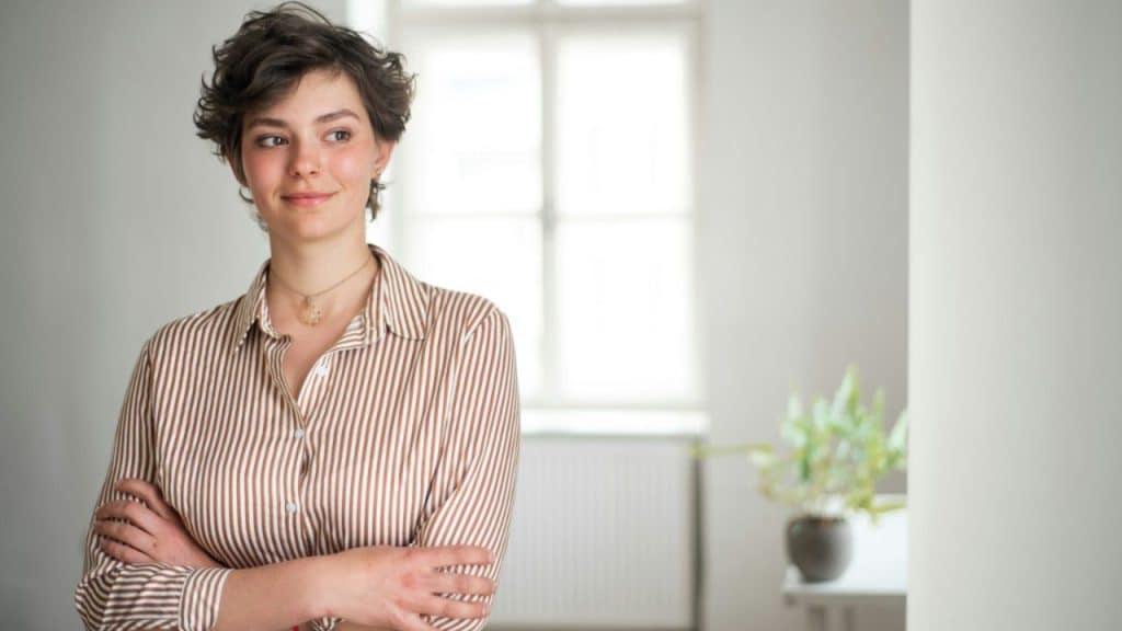 A woman with short curly hair stands with her arms crossed, looking to the side.