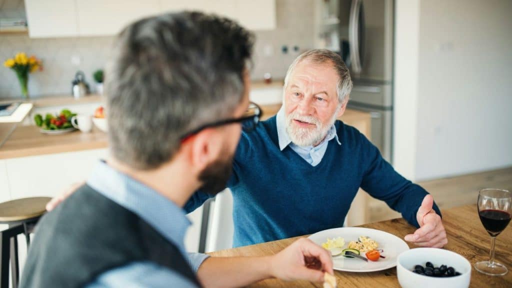 An older man talks and gestures while sitting at a dining table with another man.