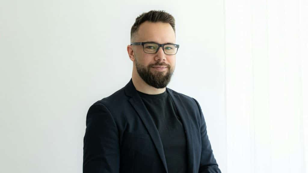 A bearded man with glasses and a dark blazer smiles slightly against a white background.