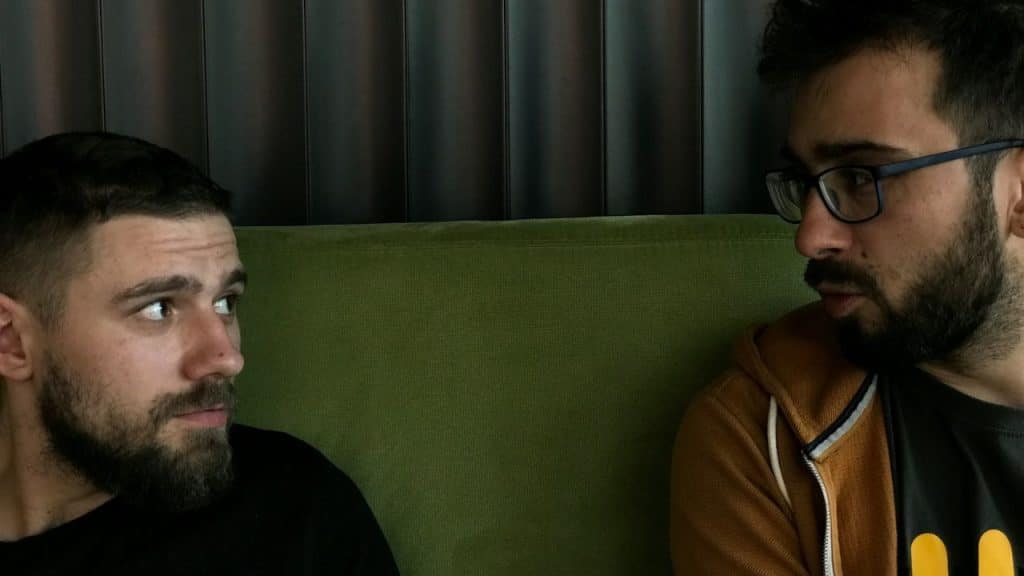 Two men with beards sit on a green couch and look at one another.