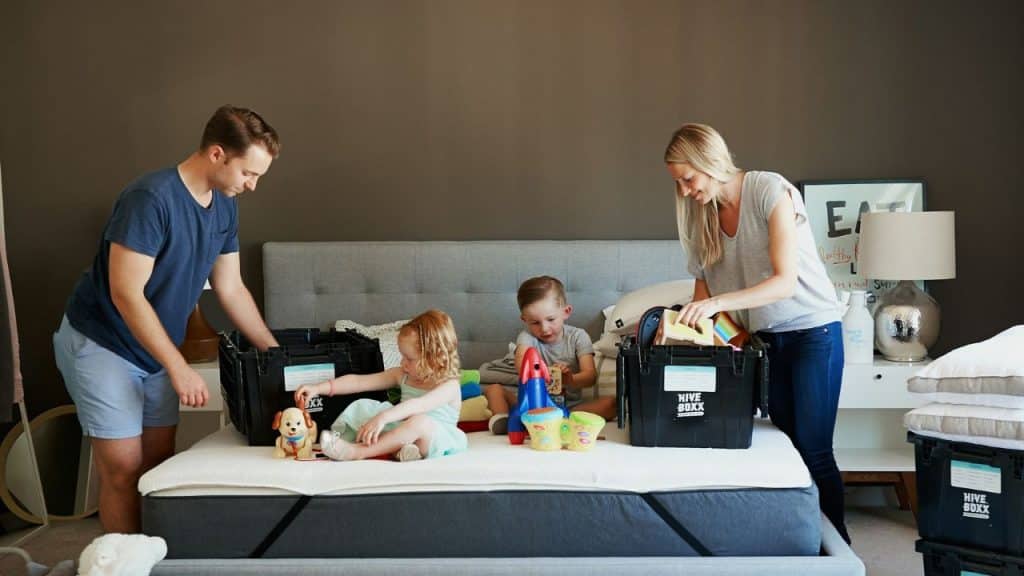 A family of four sits on a bed packing toys into large black crates.