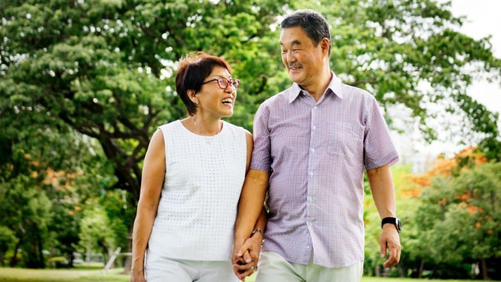 An older couple walks through a park holding hands and smiling at one another.