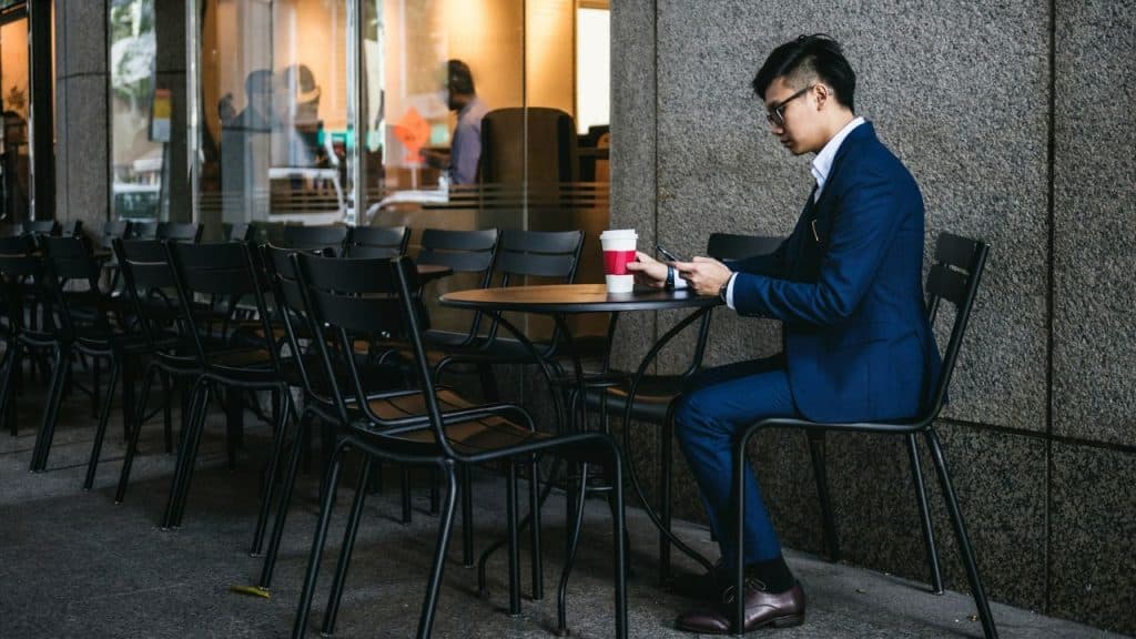 A man in a blue suit sits alone at an outdoor cafe table with coffee.