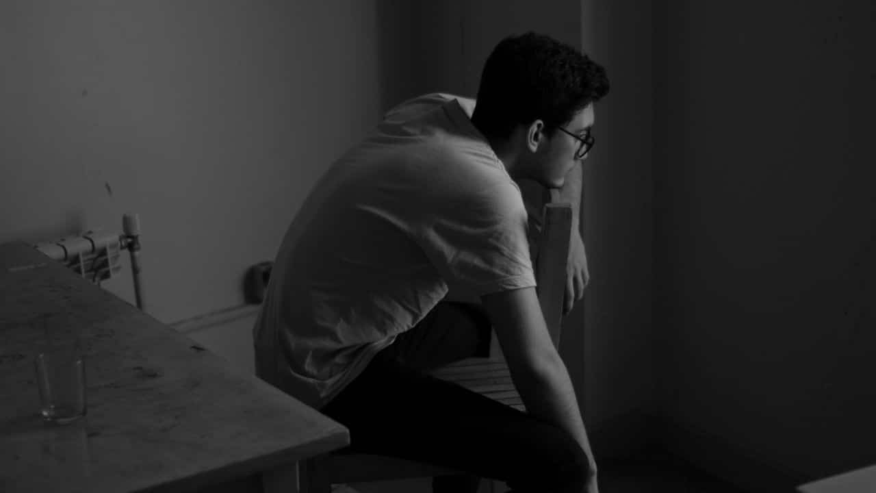 A man in a white t-shirt sits on a chair in a dark, dimly lit room.