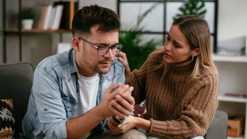 A woman comforts a distressed man by holding his hands and touching his shoulder.