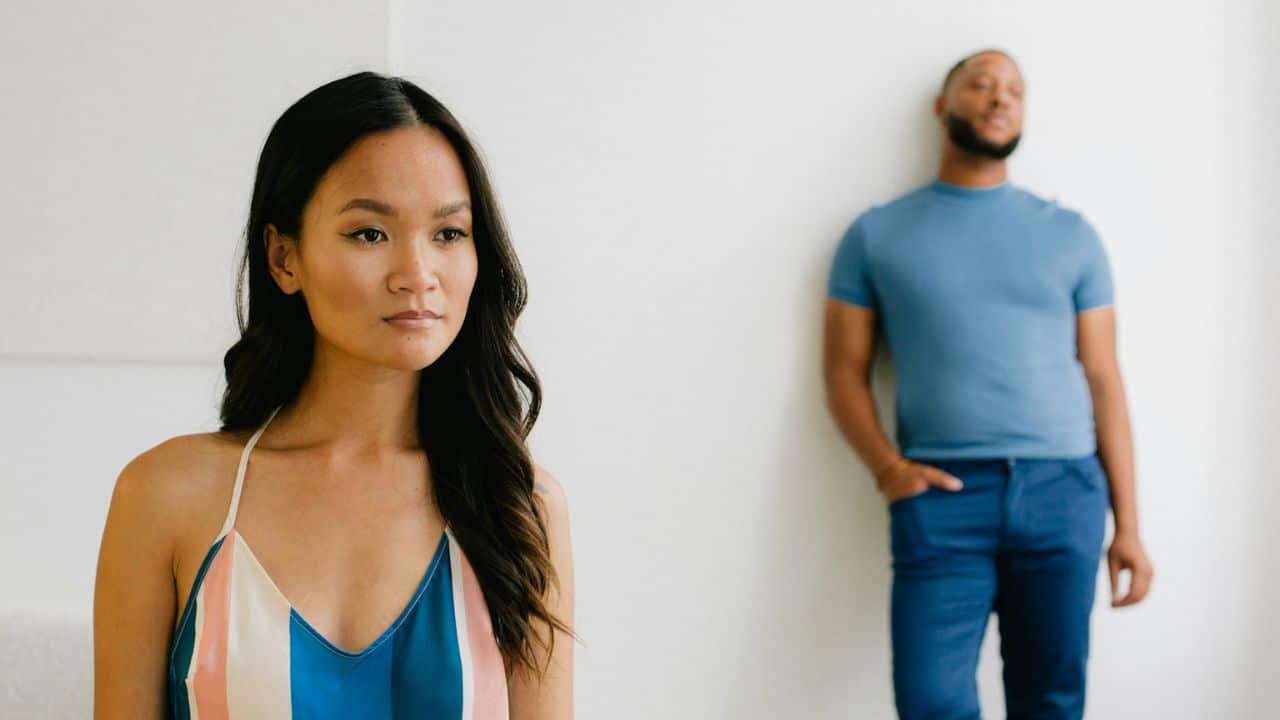 A woman in a striped top looks forward while a man stands blurred in the background.