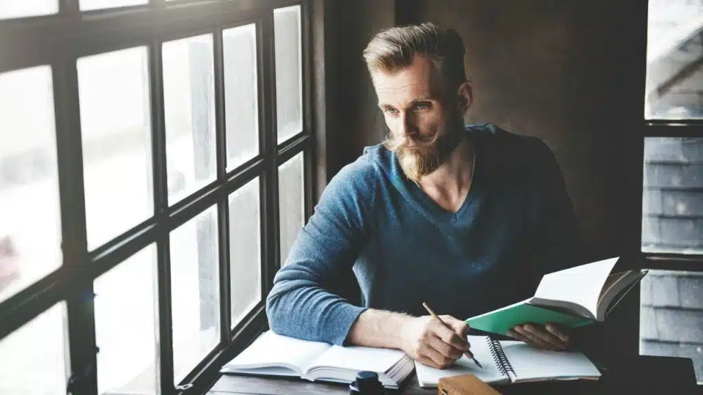 A man with a groomed beard and mustache looks out a window while writing in a notebook.