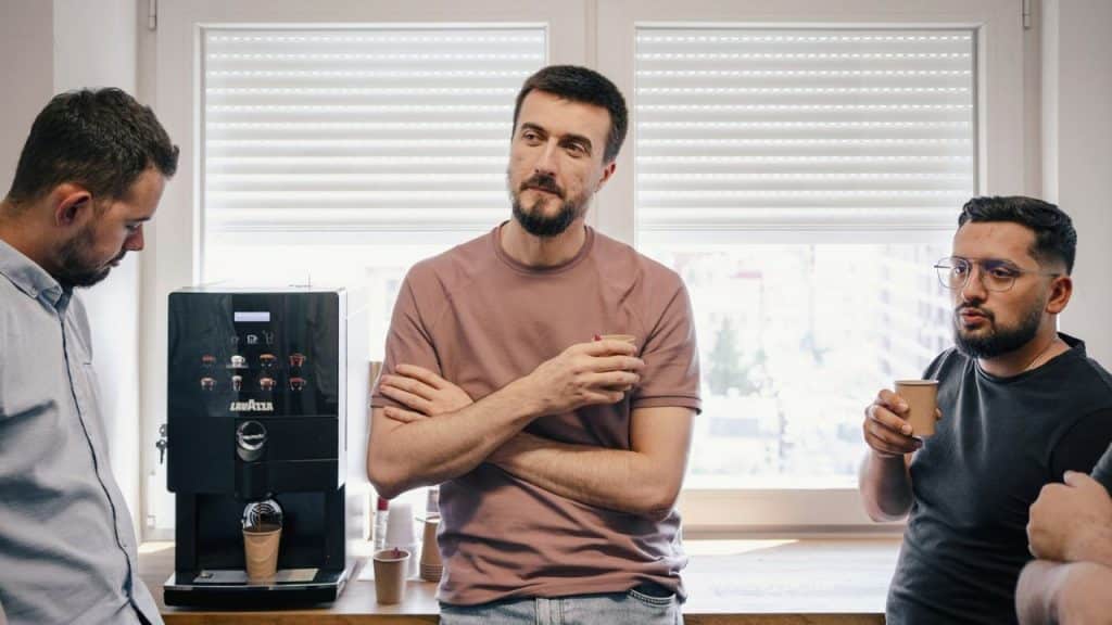 Three men stand near a coffee machine, holding cups and engaging in a serious conversation.