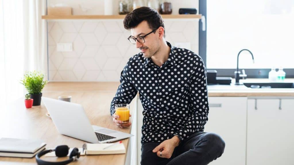 A man in a polka dot shirt sits in a kitchen looking at a laptop.