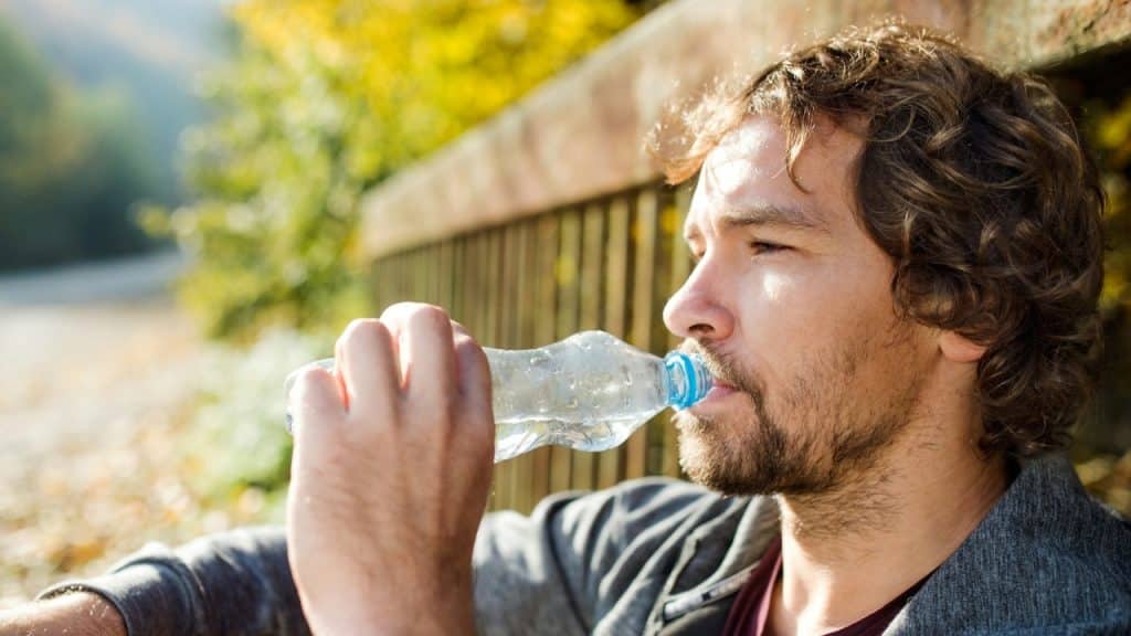 A man with curly hair and a beard drinks from a plastic water bottle outdoors.