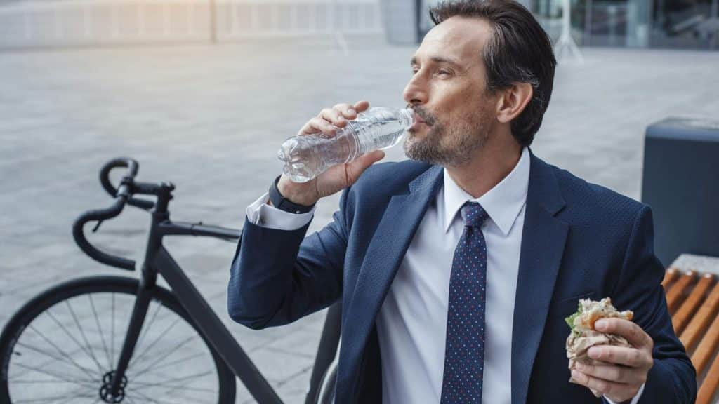 A man in a suit drinking water from a bottle while holding a sandwich.