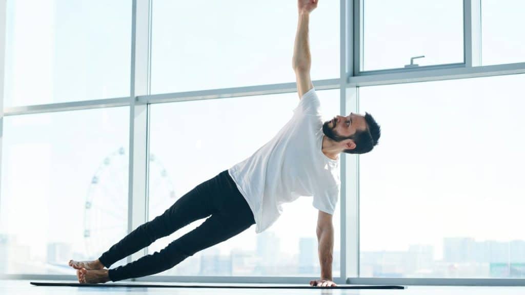 A man in a white t-shirt performs a side plank yoga pose in a studio.