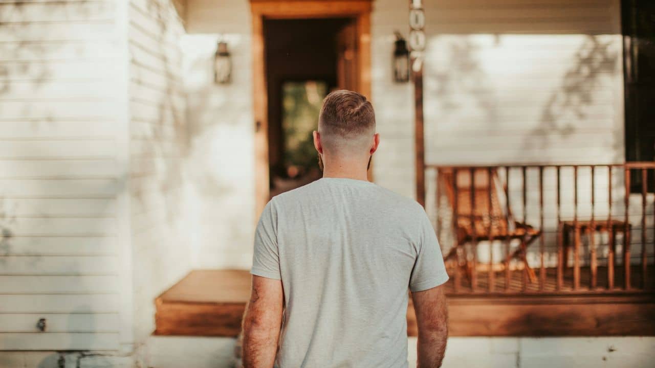 A man viewed from behind walks toward the wooden porch of a white house.
