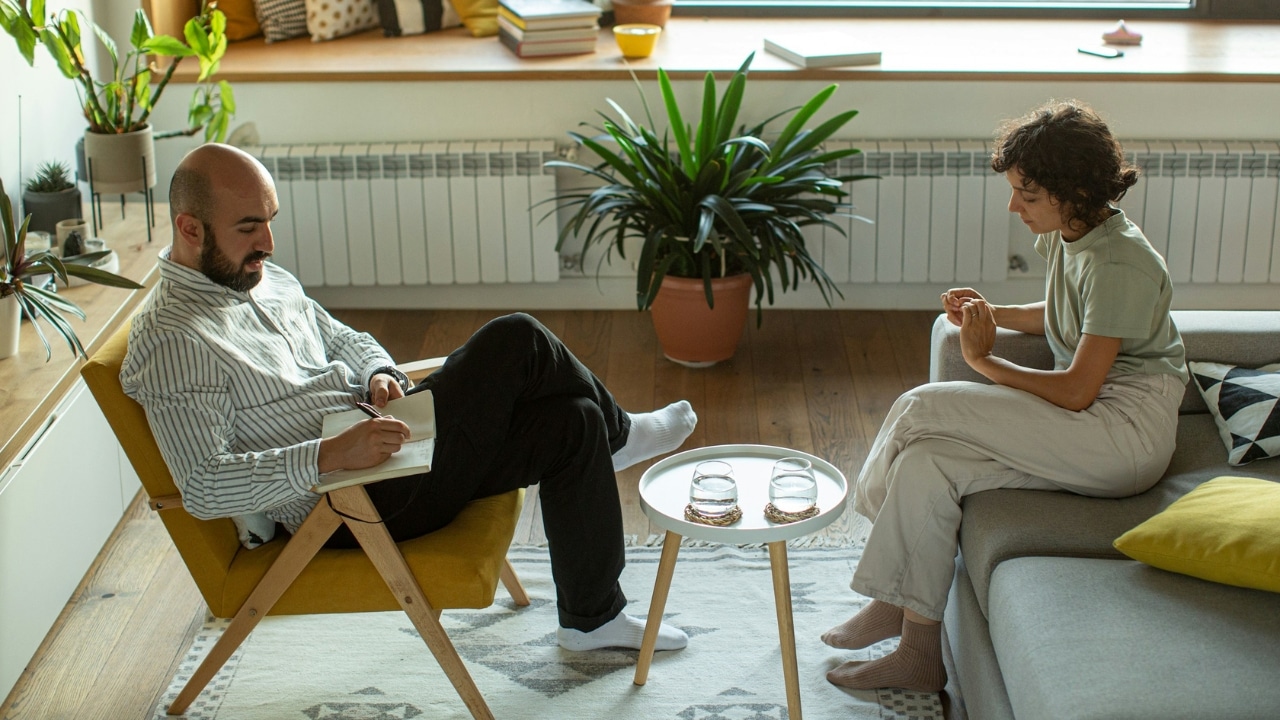 A man writes in a notebook while a woman sits opposite him in a living room.