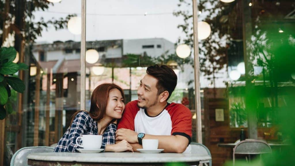 A man and woman sit at an outdoor cafe table, smiling and looking at one another.