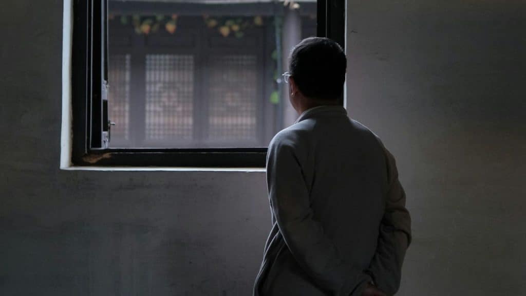 A man is viewed from behind as he stands alone, looking out of a dark window.