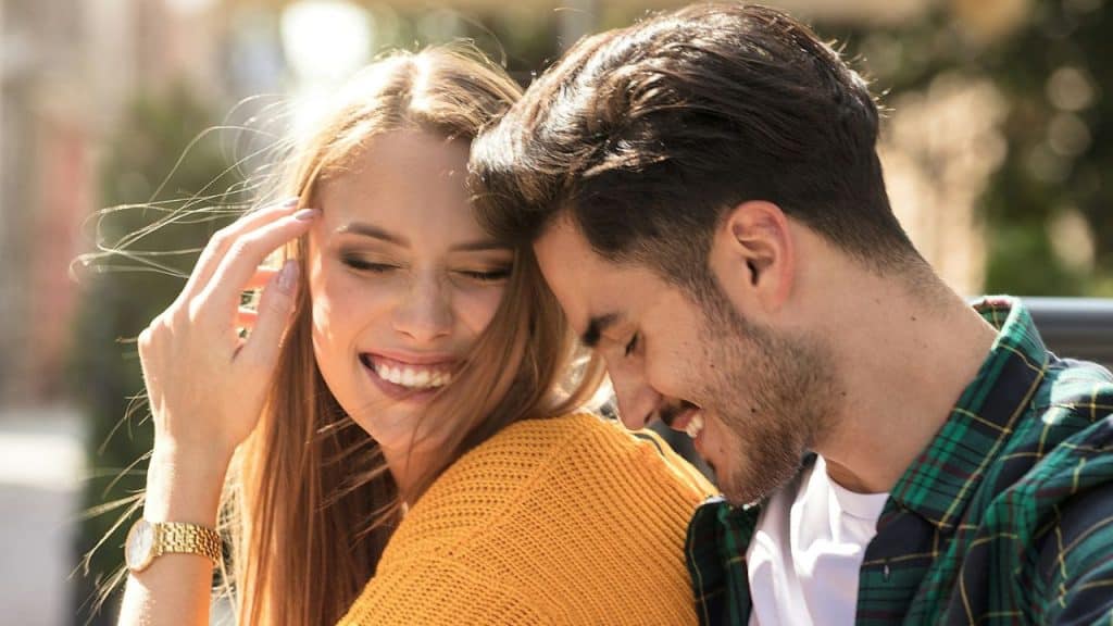 A laughing woman in a yellow top brushes her hair back while sitting next to a smiling man.