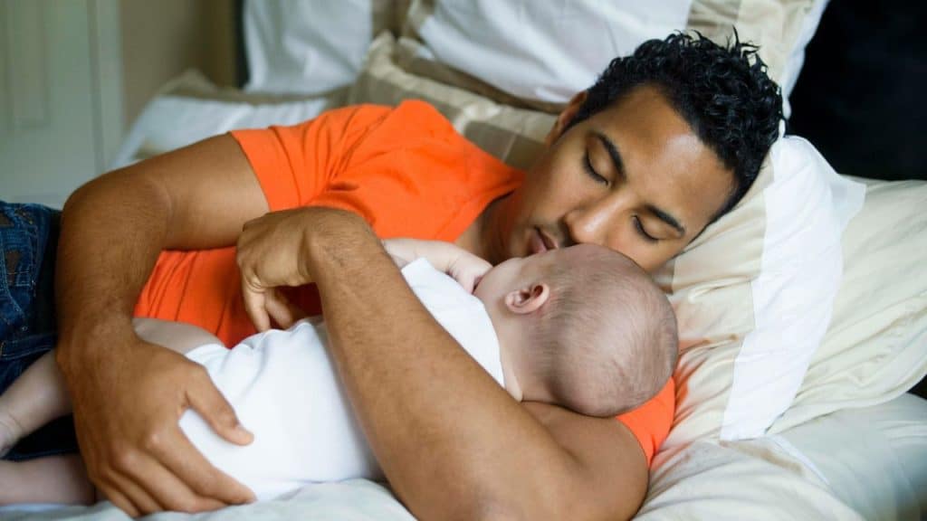 A man in an orange shirt sleeps on a bed while holding a baby.