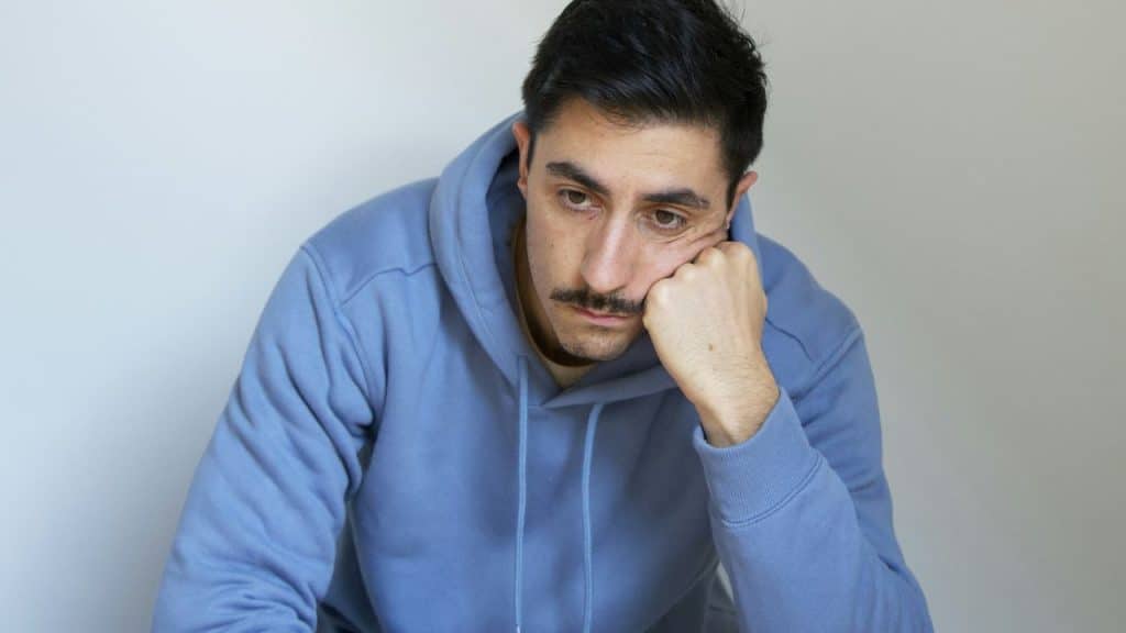 A man in a blue hoodie rests his chin on his hand with a neutral expression.