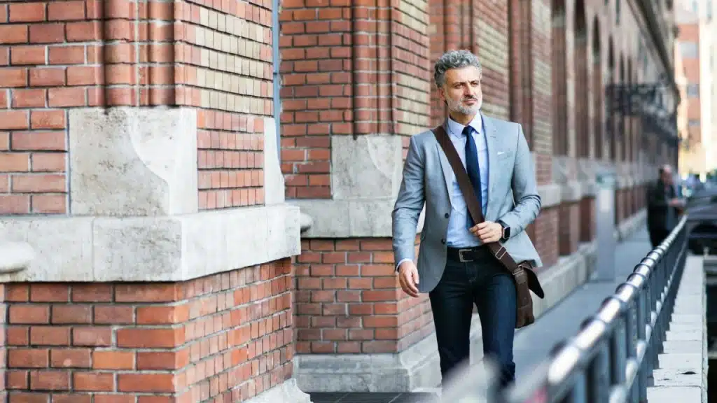 A man in a grey blazer and shoulder bag walks past a red brick building.