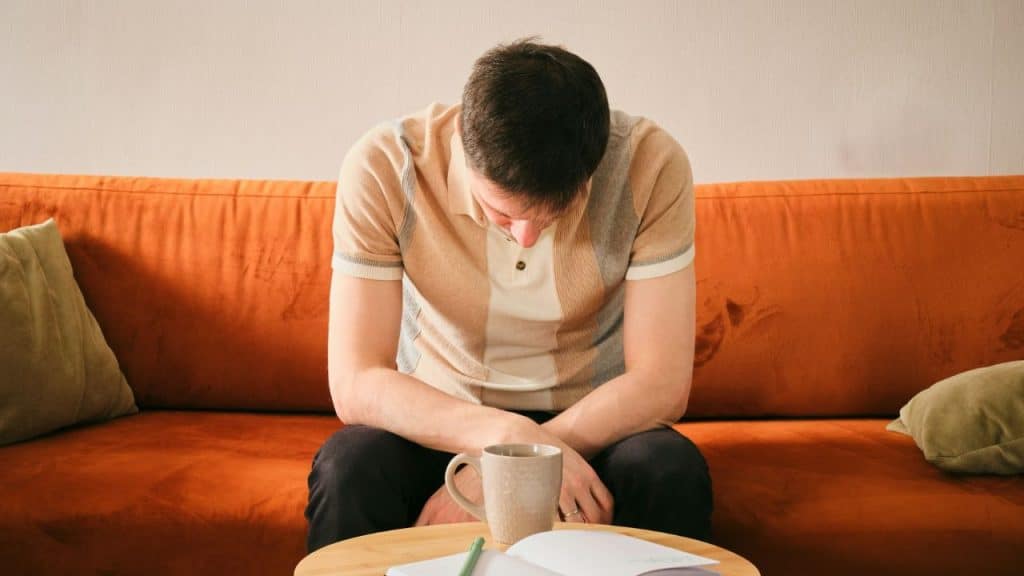 A man sits on an orange couch with his head bowed over a cup and notebook.