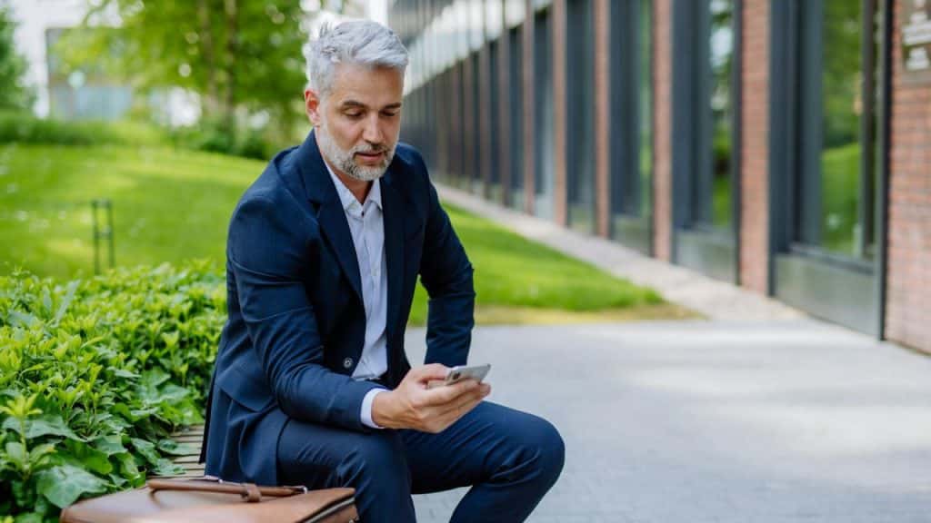 A man with grey hair in a blue suit sits outdoors and looks at a smartphone.