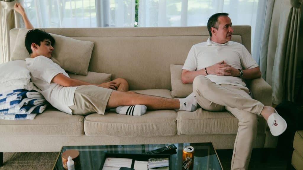 A man and a teenager sit on opposite ends of a long tan sofa.