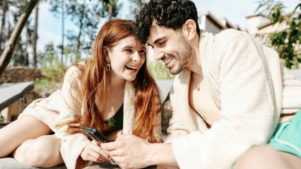 A smiling couple in robes looking at a smartphone outdoors.