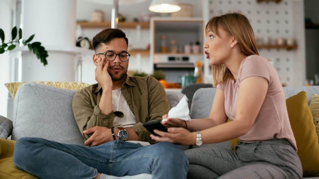 A woman talks to a man while holding a tissue and a phone on a couch.