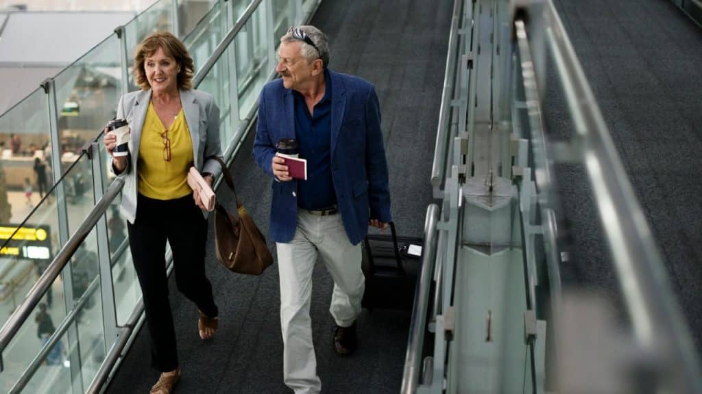 A cheerful mature couple walks up an escalator in an airport, holding coffee.