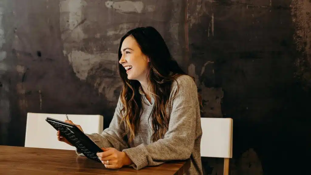 A woman smiling and holding a tablet while sitting at a table.